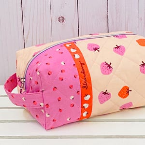 May include: A quilted cosmetic bag with a strawberry pattern. The bag has a pink handle and a zip closure. The design includes pink and orange strawberries on a light beige background, with a pink panel and the word "Strawberry".