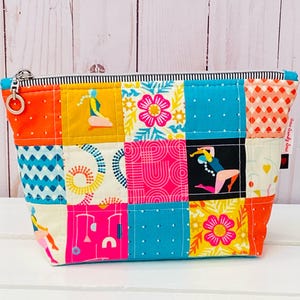Peut inclure: Trousse de maquillage patchwork colorée avec fermeture éclair. Le sac présente des carrés aux motifs variés en orange, bleu, rose et jaune, avec des motifs floraux et géométriques. Le haut est rayé et le curseur de la fermeture est argenté.
