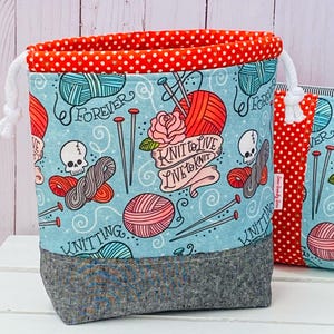 Peut inclure: Un sac de projet de tricot avec un fond bleu clair présentant un motif répété de pelotes de laine, d'aiguilles, de crânes et les mots "Forever" et "Knit to Live, Live to Knit". Le haut est rouge à pois blancs, et le bas est gris.