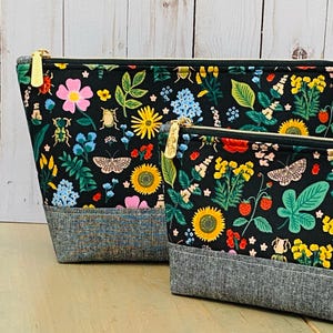 May include: Two black zippered pouches with a floral and insect pattern. The design includes pink, yellow, and blue flowers, butterflies, and beetles. The bottom of the pouches is a grey tweed fabric. Gold zippers.