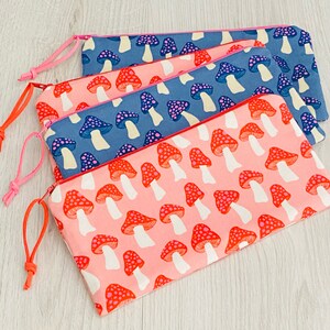 Peut inclure: Trois pochettes zippées avec des motifs de champignons. La pochette du haut est bleue avec des champignons blancs, la pochette du milieu est bleue avec des champignons blancs et roses, et la pochette du bas est rose avec des champignons rouges et blancs.