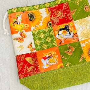 May include: A drawstring pouch featuring a patchwork design of autumn-themed fabric squares. The pattern includes sunflowers, orange butterflies, red leaves, and illustrations of cats in baskets and pumpkins. The base is a solid textured lime green fabric.