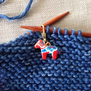 May include: A blue knitted fabric with a red and blue horse-shaped stitch marker attached to a wooden knitting needle.