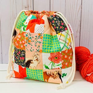 May include: A colourful patchwork drawstring bag featuring floral, horse, and geometric patterns in orange, green, pink, and black. The bag has a cream drawstring closure and the text "Sew Sandy Sew" is visible. A ball of orange yarn is next to the bag.