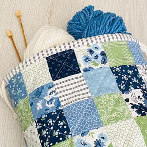 Peut inclure: Un sac de projet de tricot en tissu patchwork dans les tons de bleu et de vert, rempli de fil blanc et bleu et d'aiguilles à tricoter en bois. Le sac a une fermeture à cordon et une bordure rayée.
