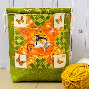 May include: A drawstring fabric bag featuring a quilted patchwork design with a central orange square depicting a black cat in a wheelbarrow surrounded by sunflowers. The bag has a green border with butterfly prints and cream-coloured drawstrings.