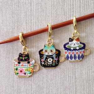 May include: Three gold cat stitch markers with colourful teacup designs. Each marker features a different cat with a unique design and colour scheme. The markers are attached to a wooden knitting needle.