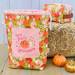 May include: A fabric drawstring bag and a matching rectangular zippered pouch, both featuring a green background with a pattern of orange and pink pumpkins and white flowers. The bag has a pink square patch with the text hey there pumpkin.