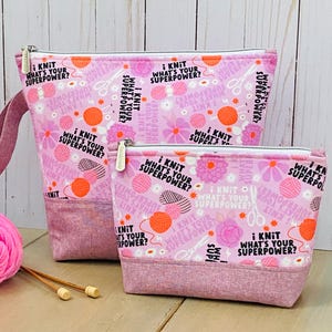 May include: Two pink zippered pouches with a floral and yarn ball pattern. The text "i knit what's your superpower?" is printed on the pouches. The bottom of the pouches are a darker pink fabric. Knitting needles and yarn are in the foreground.