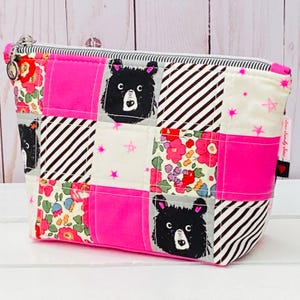 Peut inclure: Une trousse de maquillage patchwork avec fermeture éclair. Le sac présente des tissus roses, floraux, à rayures noires et blanches et des visages d'ours. Il a une doublure rayée et une fermeture éclair argentée.