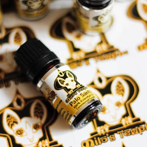 May include: A small, dark brown glass bottle with a black cap and a white label. The label has a black and gold illustration of a cat and the text "Milo's Temple Palo Santo 100% Essential Oil Ceremonial Grade".