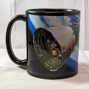 Puede incluir: Taza de café de cerámica negra con asa blanca. La taza presenta una imagen colorida de una nave espacial en órbita alrededor de la Tierra.