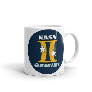 Puede incluir: Taza de cerámica blanca con el logotipo de NASA Gemini. El logotipo es un círculo azul marino con la palabra "NASA" encima de un símbolo "II" dorado, flanqueado por dos estrellas blancas y la palabra "GEMINI" debajo.