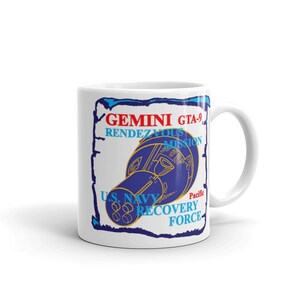 Puede incluir: Taza de cerámica blanca con una ilustración azul y dorada de la Misión Gemini GTA-9 Rendezvous. La taza también incluye el texto "U.S. Navy Recovery Force" en azul y dorado.