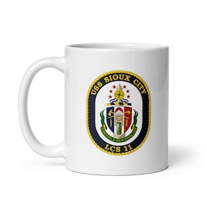 Puede incluir: Taza de café de cerámica blanca con un escudo azul marino, dorado y rojo con el texto "USS SIOUX CITY" y "LCS 11". El escudo también incluye un escudo con un guerrero nativo americano y el texto "FORGING A NEW FRONTIER".