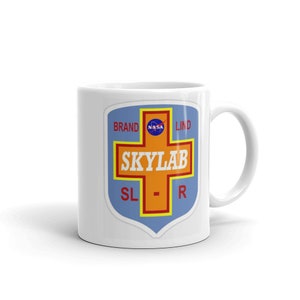 May include: White ceramic mug with a blue and orange Skylab logo. The logo features a red cross with the words "Skylab" in white letters. The words "Brand", "Lind", and "NASA" are also visible on the logo.