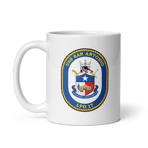May include: White ceramic mug featuring the USS San Antonio emblem. The emblem is a blue oval with a gold rope border, displaying a cannon, star, and text. The text "USS SAN ANTONIO" and "LPD 17" are visible.