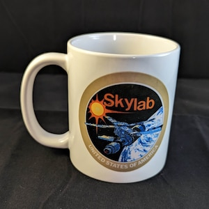 May include: White ceramic mug with a graphic of the Skylab space station orbiting Earth. The graphic is surrounded by a gold circle with the text "Skylab" and "United States of America".