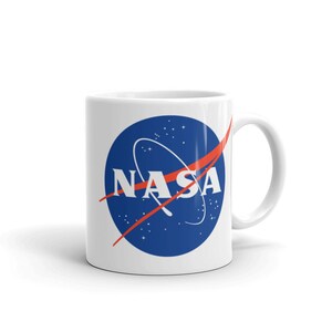 May include: White ceramic mug with a blue and red NASA logo. The logo features a blue circle with a red curved line and white stars. The word "NASA" is written in white letters inside the circle.
