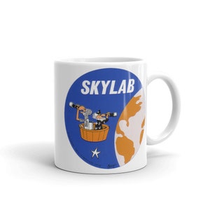 Könnte beinhalten: Eine weiße Keramiktasse mit einem blauen und orangefarbenen Skylab-Logo. Das Logo zeigt eine Karikatur von zwei Astronauten in einer Raumkapsel, die um eine stilisierte Erde kreisen. Der Text "SKYLAB" steht in großen, fetten Buchstaben über den Astronauten. Der Text "UNITED STATES  SYNDICATE 1970" steht in kleineren Buchstaben unter den Astronauten.
