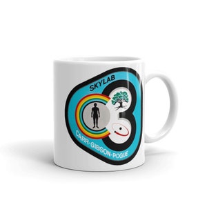May include: White ceramic mug with a colorful Skylab mission patch. The patch features a blue background with a rainbow, a silhouette of a person, a tree, and a red ring. The text "SKYLAB" and "CARR-GIBSON-POGUE" are printed on the patch.