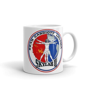 Könnte beinhalten: Weiße Keramiktasse mit einer bunten Grafik eines Mannes in einem Kreis. Der Kreis trägt den Text "BEAN GARRIOTT LOUSMA" und "SKYLAB II" um den Außenrand.