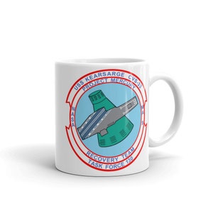 May include: White ceramic mug with a red, white, and blue circular design. The design features a stylized image of a spacecraft being recovered by a ship. The text "USS KEARSARGE CVS-33 PROJECT MERCURY RECOVERY TEAM TASK FORCE 130" is printed around the image.