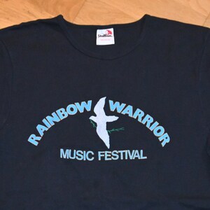 May include: A black t-shirt with a blue and green graphic of a dove with an olive branch in its beak. The graphic is arched over the text "Rainbow Warrior Music Festival".