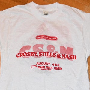 Könnte beinhalten: Weißes T-Shirt mit rotem Druck, das für ein Konzert von Crosby, Stills & Nash im Mann Music Center in Philadelphia am 4. und 5. August wirbt. Der Text "electric factory concerts" ist ebenfalls auf dem Shirt gedruckt.