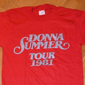 May include: A red t-shirt featuring the text "DONNA SUMMER TOUR 1981" in a silver, stylized font. The shirt is a classic crew neck style, likely made of cotton or a similar fabric. The vintage design is centered on the front.