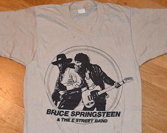 May include: A beige t-shirt with a black and white image of Bruce Springsteen and the E Street Band. The image is in a circle with the band's name printed below.