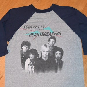 May include: A gray and navy blue baseball tee shirt with a black and white photo of Tom Petty and the Heartbreakers. The text "Tom Petty and the Heartbreakers" is printed in white on the shirt.
