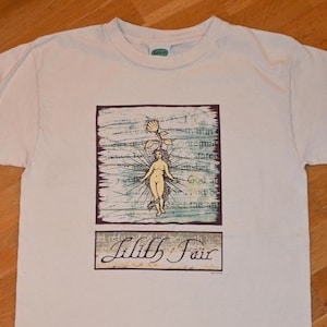May include: A white short-sleeve t-shirt featuring a central graphic print. The design includes a stylized figure with a flower head, radiating lines, and text overlays. Below the graphic, the text Lilith Fair is printed in a script font.