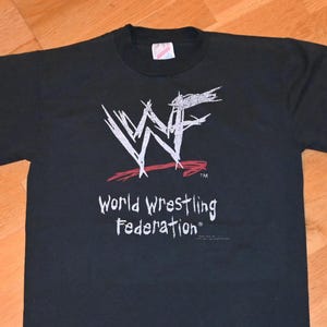 Puede incluir: Camiseta negra con el logotipo de la World Wrestling Federation en blanco y rojo. El texto "World Wrestling Federation" está impreso debajo del logotipo en blanco. La camiseta es de material suave.