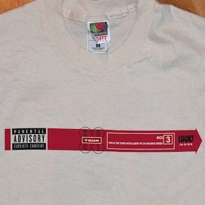 Puede incluir: Una camiseta blanca con una etiqueta de advertencia para padres roja y negra y el texto "2001 Tour" y "Look! Inside" con una flecha roja. El texto "This is the third installment of an ongoing series" también es visible.