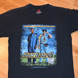 Puede incluir: Camiseta negra con el póster de la película "Half Baked". El diseño muestra a dos hombres frente a un fondo azul y verde, el título de la película en amarillo y el texto "Keep Off The Grass".