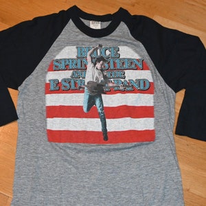 May include: Gray raglan-style t-shirt with black sleeves. The front features a graphic of Bruce Springsteen and the E Street Band, with a photo of Springsteen in front of a red and white striped background.