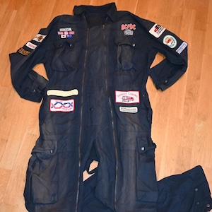 May include: A dark blue jumpsuit with multiple patches, including a "Brodkum" patch, an "AC/DC" patch, and a "Southern Counties" patch. The jumpsuit has two large cargo pockets on the legs.