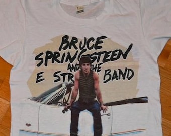 May include: White t-shirt featuring a graphic of Bruce Springsteen and the E Street Band. The graphic shows Springsteen sitting on a car. The text "BRUCE SPRINGSTEEN and THE E STREET BAND" is printed above the image.