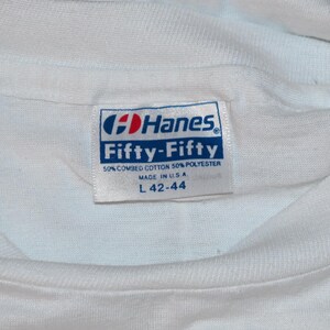 May include: A white t-shirt featuring a rectangular Hanes Fifty-Fifty brand label sewn into the inner neckline. The label displays the text Hanes, Fifty-Fifty, 50% combed cotton 50% polyester, Made in U.S.A., and size L 42-44 in blue and red ink.