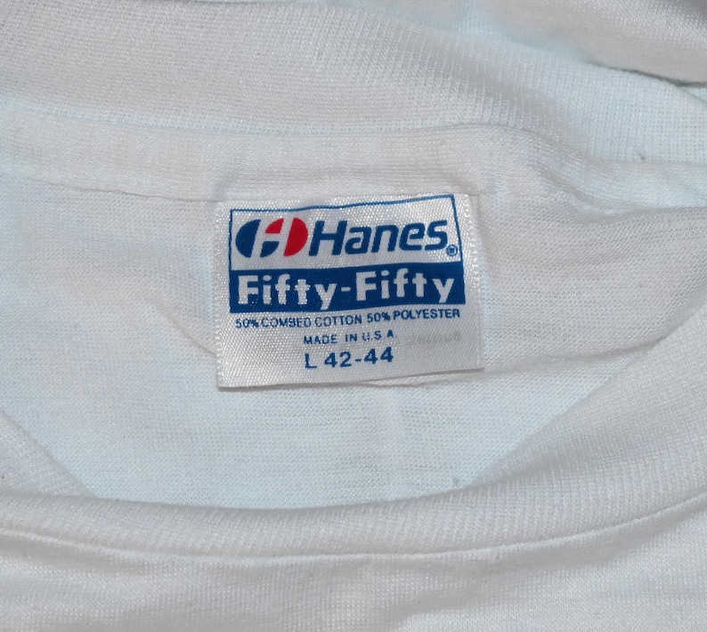 May include: A white t-shirt featuring a rectangular Hanes Fifty-Fifty brand label sewn into the inner neckline. The label displays the text Hanes, Fifty-Fifty, 50% combed cotton 50% polyester, Made in U.S.A., and size L 42-44 in blue and red ink.