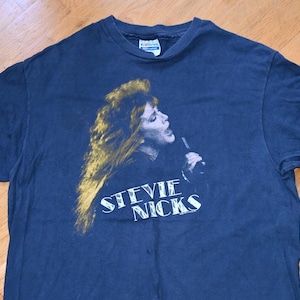 May include: A dark blue short-sleeved t-shirt featuring a graphic print of Stevie Nicks singing into a microphone. The print displays her with long, flowing hair in shades of gold and brown, with the text STEVIE NICKS printed in white below.