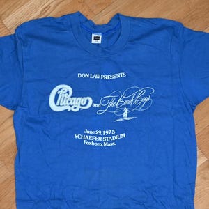 May include: A blue t-shirt featuring the text "Chicago and The Beach Boys" in white script. The shirt also includes the date "June 29, 1975" and the location "Schaefer Stadium Foxboro, Mass."
