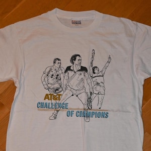 May include: A white short-sleeve t-shirt featuring a black line-art graphic of three tennis players in action. The text AT&T CHALLENGE OF CHAMPIONS is printed in blue and gold below the figures. The shirt has a crew neckline and a tag at the collar.