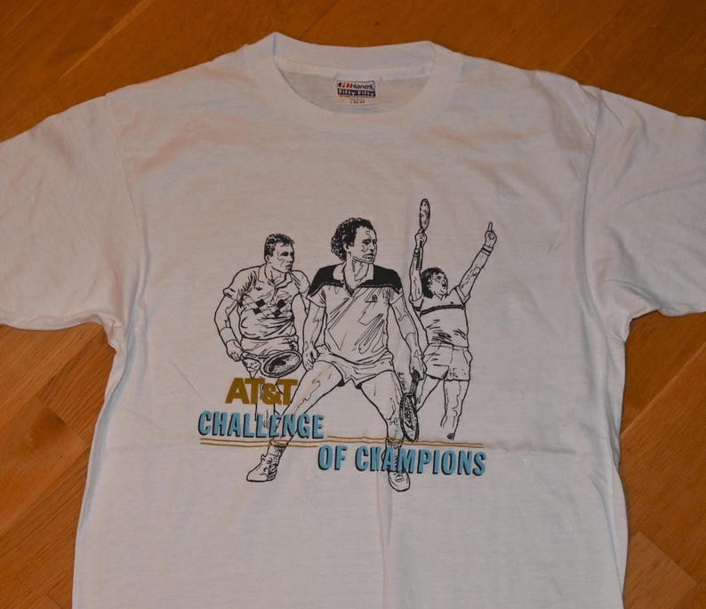 May include: A white short-sleeve t-shirt featuring a black line-art graphic of three tennis players in action. The text AT&T CHALLENGE OF CHAMPIONS is printed in blue and gold below the figures. The shirt has a crew neckline and a tag at the collar.