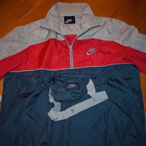 May include: A gray, red, and navy blue Nike anorak jacket. The jacket has a half-zip closure, a front pocket with a flap, and the Nike logo on the chest and pocket. The jacket is made of a lightweight material.