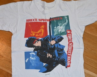 May include: White t-shirt featuring a graphic of Bruce Springsteen and The Street Band. The design includes images of Springsteen playing guitar, set against red, green, and blue backgrounds. The band's name is printed vertically in red.