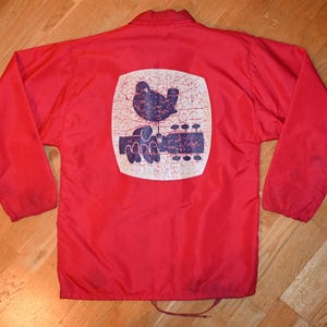 Op de afbeelding: Rode nylon jas met een trekkoordzoom. De achterkant heeft een verweerd witte vierkant met een blauwe afbeelding van een vogel, een hand en een gitaar. De jas heeft lange mouwen en een kraag.