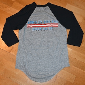 May include: A gray raglan baseball shirt with black sleeves. The back of the shirt features the text "BORN IN THE USA TOUR '84-'85" in blue and red lettering, reminiscent of the American flag.