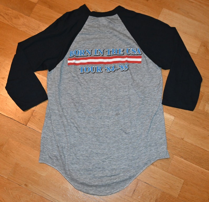 May include: A gray raglan baseball shirt with black sleeves. The back of the shirt features the text "BORN IN THE USA TOUR '84-'85" in blue and red lettering, reminiscent of the American flag.